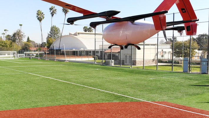 A white plane with multiple rotors and reddish wing and tail parts flies in front of Caltech's Brown Gym.