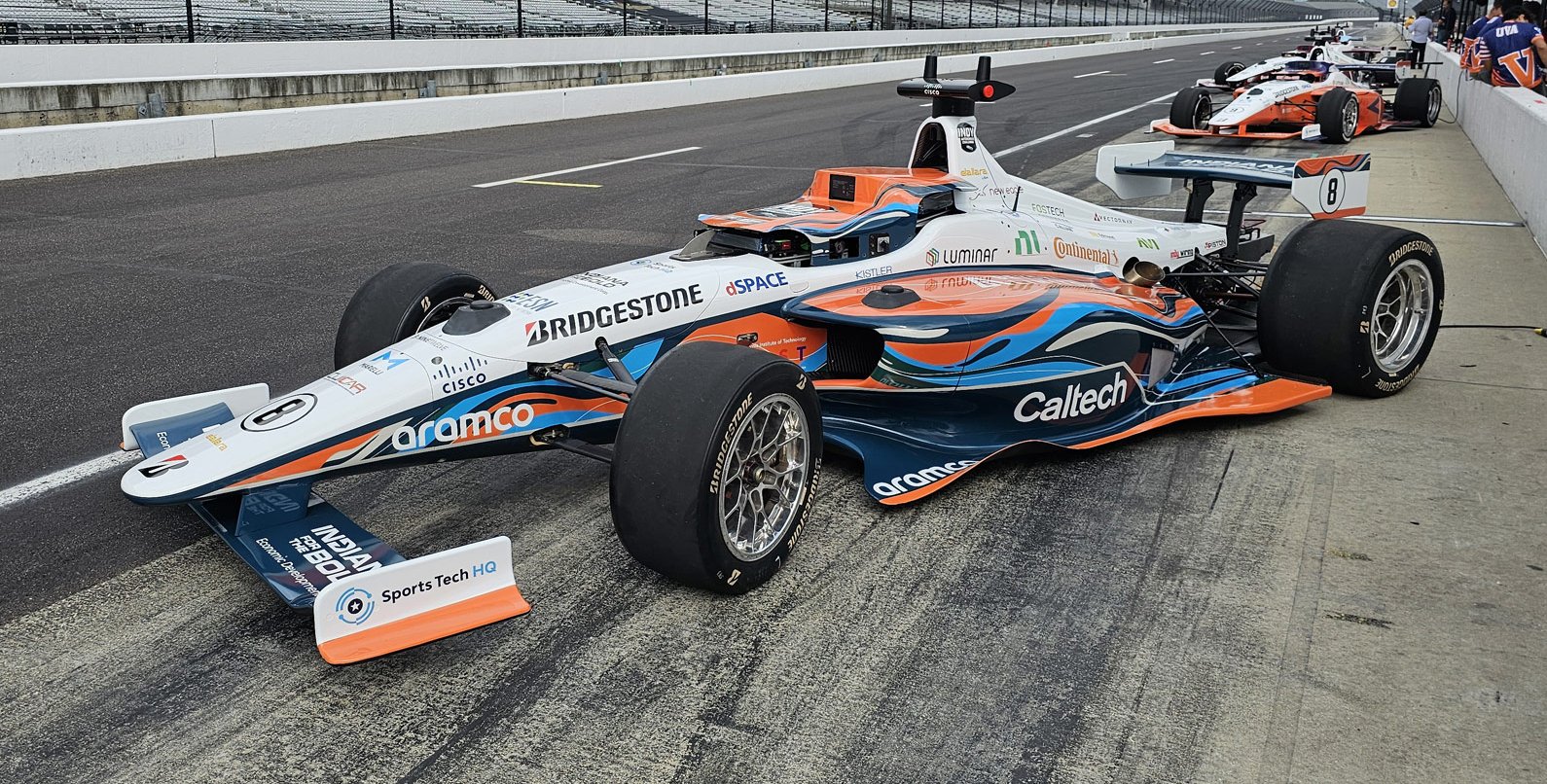 an orange, white, and light blue race car with a Caltech label parked on a racetrack with several other race cars in the background