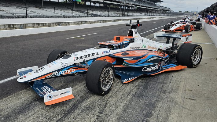 an orange, white, and light blue race car with a Caltech label parked on a racetrack with several other race cars in the background