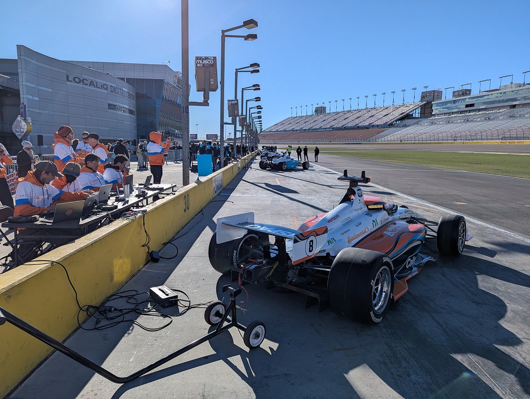 Caltech's autonomous racecar parked in pit lane of a raceway with Caltech team members working on laptops to get the car ready to race