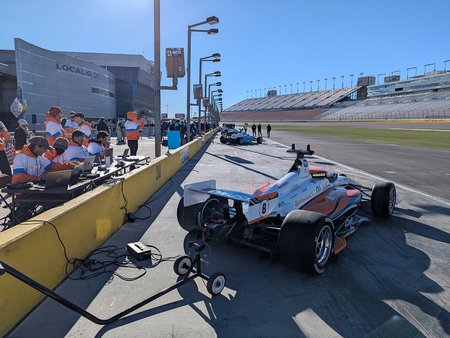 Caltech's autonomous racecar parked in pit lane of a raceway with Caltech team members working on laptops to get the car ready to race