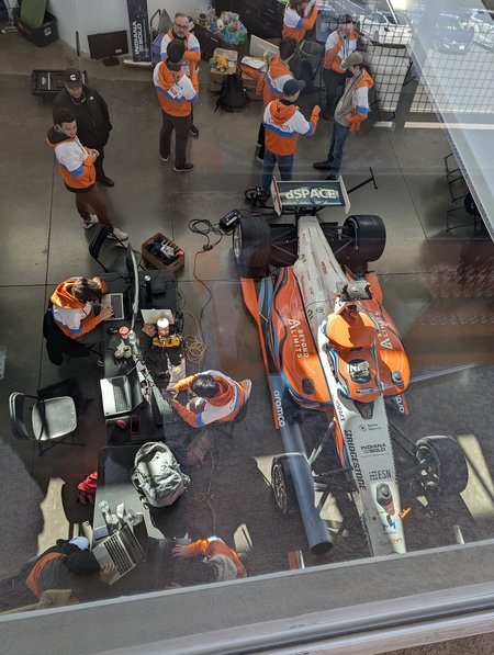 an overhead view of Caltech's autonomous race car and about a dozen members of the team that support it wearing orange, blue, and white Caltech jackets