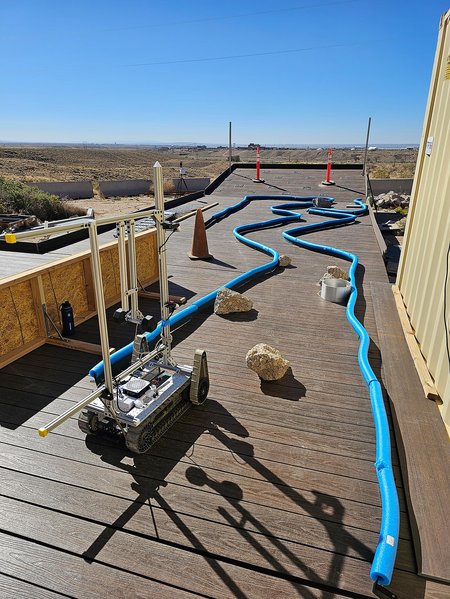 A tracked vehicle making its way through a track set up on a wooden walkway with blue pool noodles outlining the curves of the track.
