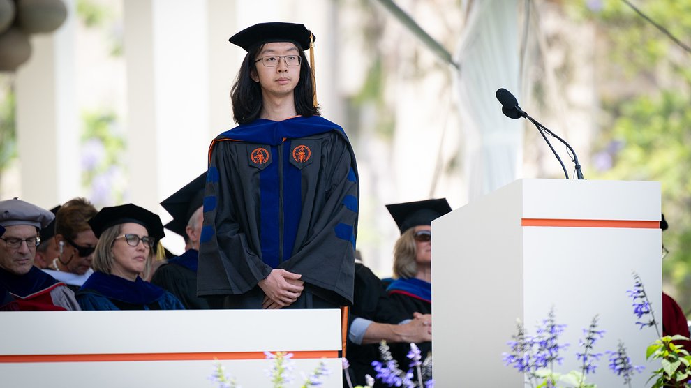 A young man with long dark hair and glasses dressed in his commencement robe stands as a commendation for him is read out.