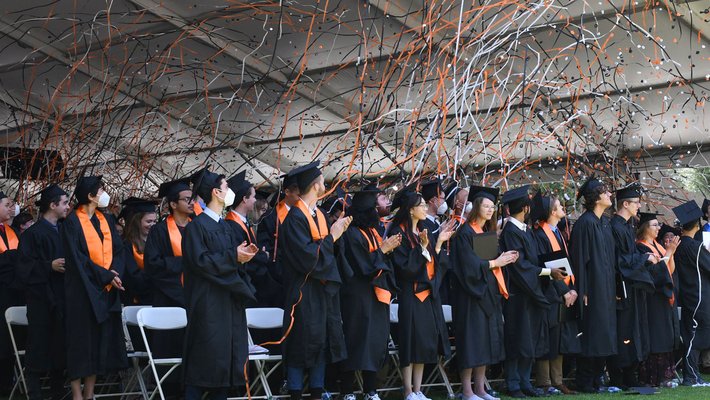 Students in caps and gowns celebrate commencement as orange confetti flies in the air.