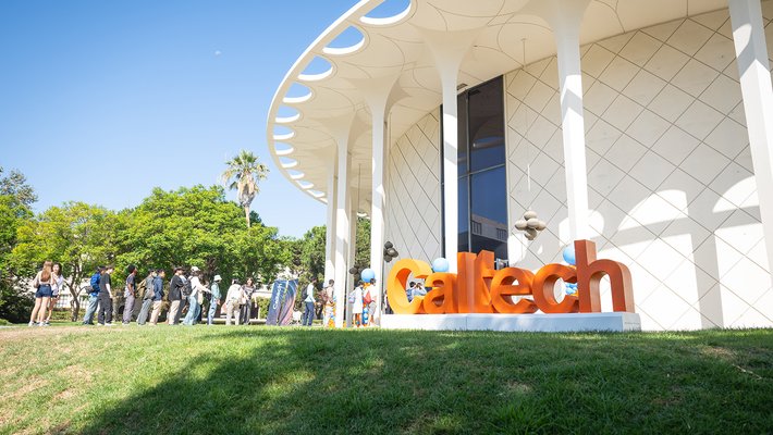 Students walk into the Beckman Auditorium. In the foreground are large orange letters spelling out "Caltech."