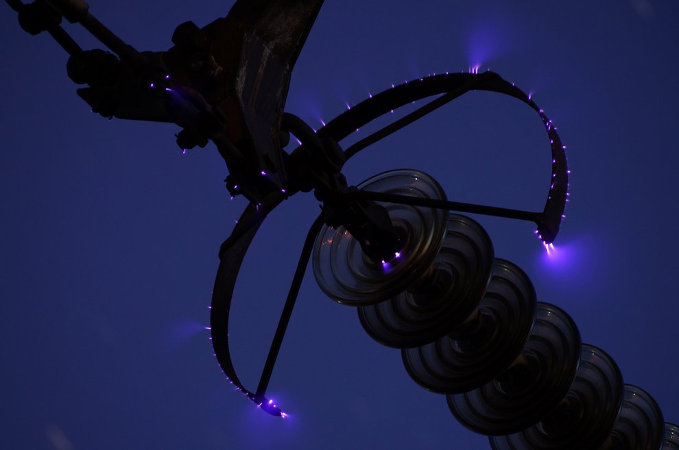 A night-time photograph of a high-voltage power line. Softly feathered electricity leaks off the line's surfaces into the air.