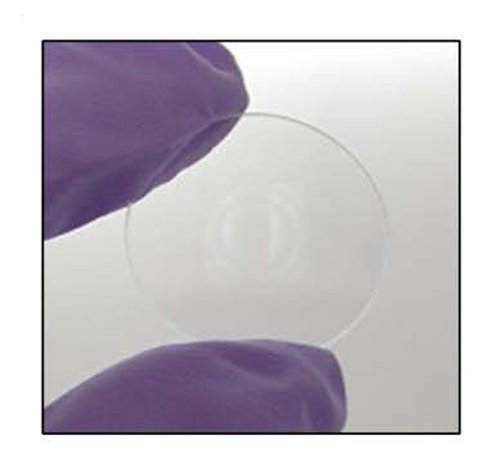 Fingers wearing a rubber glove appear in this close-up image of a diffractive optical element. The DOE appears as a small glass disc with a paint pattern at its center.