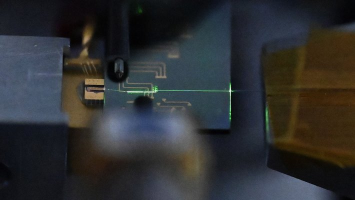 A small circuit board appears in dim lighting. A green laser beam emerges from it.