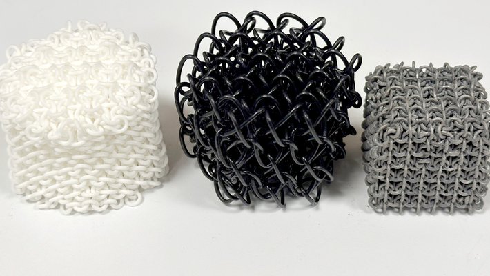 Three polycatenated architected materials printed out in three different materials: nylon, TPU, and steel. The nylon PAM is white and cylindrical; the TPU one is black and roughly spherical; the steel one is dark gray and cubical.