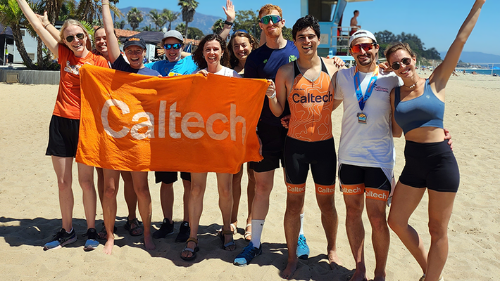 ten people standing on the beach holding an orange towel that says Caltech