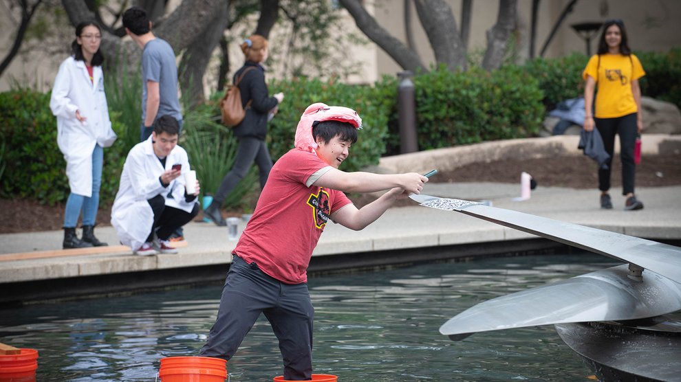 Members of the "God Damn Dino Gang" stack cross Caltech Hall Pond using plastic buckets and wooden beams in order to scan a QR code.