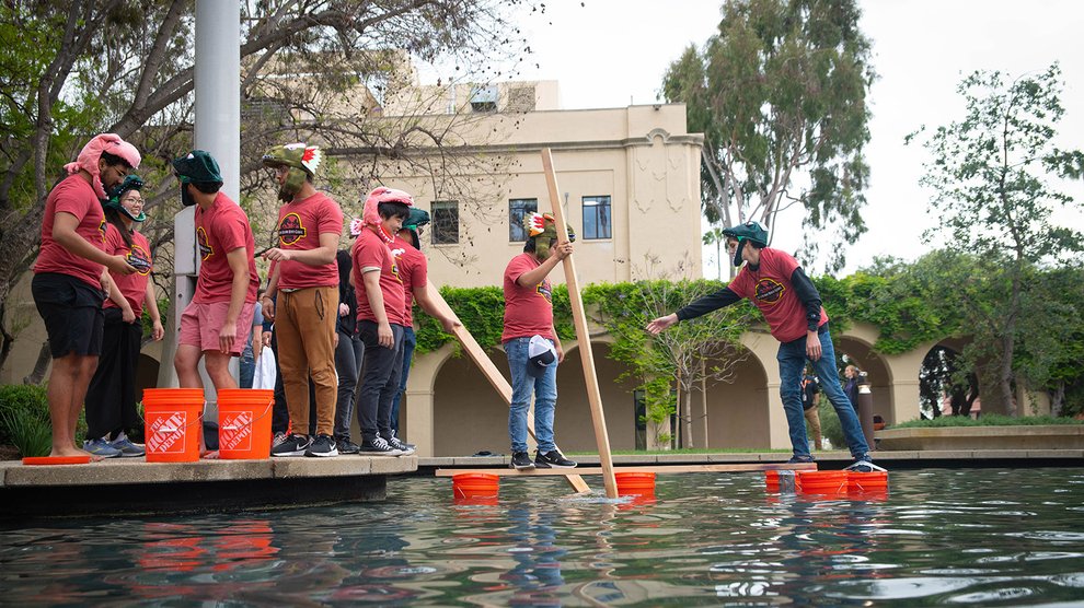 Members of the "God Damn Dino Gang" stack cross Caltech Hall Pond using plastic buckets and wooden beams in order to scan a QR code.