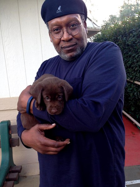 Don Crewell, former director of financial aid, holds a chocolate lab puppy and looks into the camera. He wears a beret.