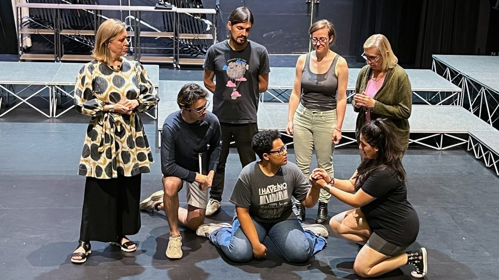 A group of six people surround and look at Maria Azcona Baez, who is sitting on the floor of a stage.