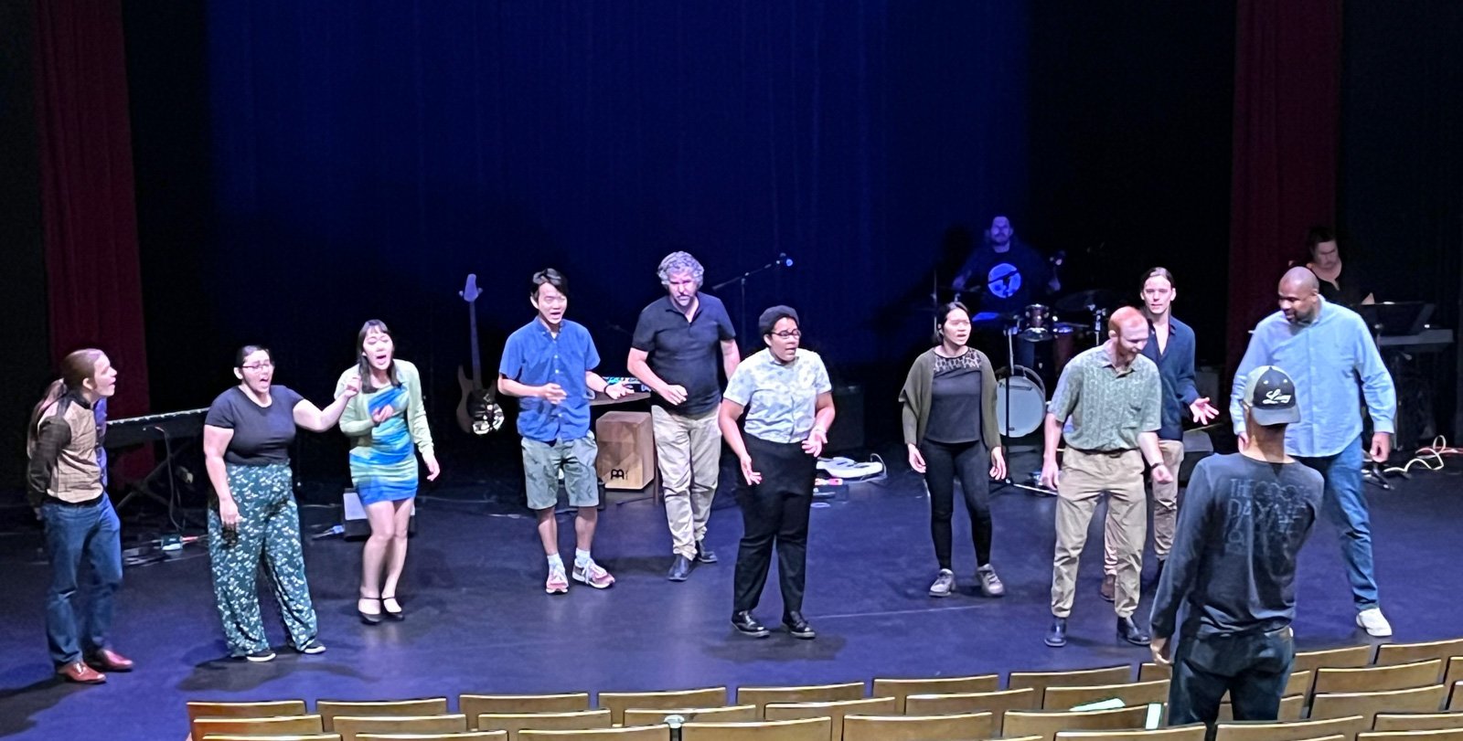 A group of about 10 people perform on a stage and Brian Brophy stands in the audience and directs them