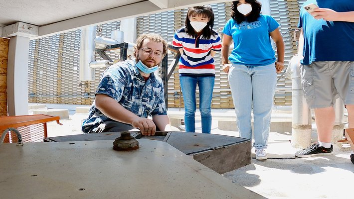 three people watch as one kneels and touches a large wedge-shaped object