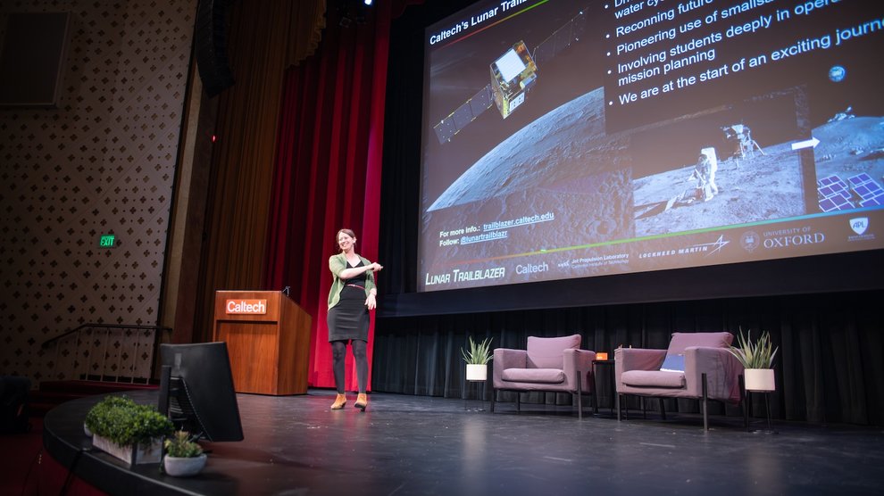 A speaker smiles and gestures onstage in front of a slide featuring the Lunar Trailblazer mission