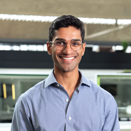 Eshaan Patheria wears round glasses and a checked blue button-up shirt and smiles at the camera. A modern urban structure is behind him.