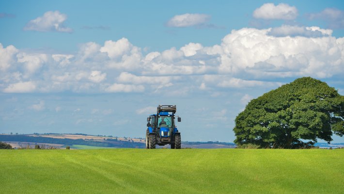 A tractor moves across a low, rolling hilld of green grass. A sky with puffy clouds is overhead.