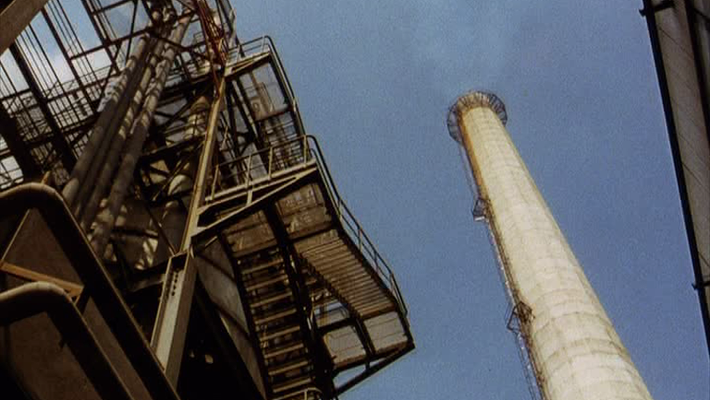 A grainy still from a vintage film. The camera points upward through the infrastructure of an oil refinery, showing a smokestack set against a blue sky.