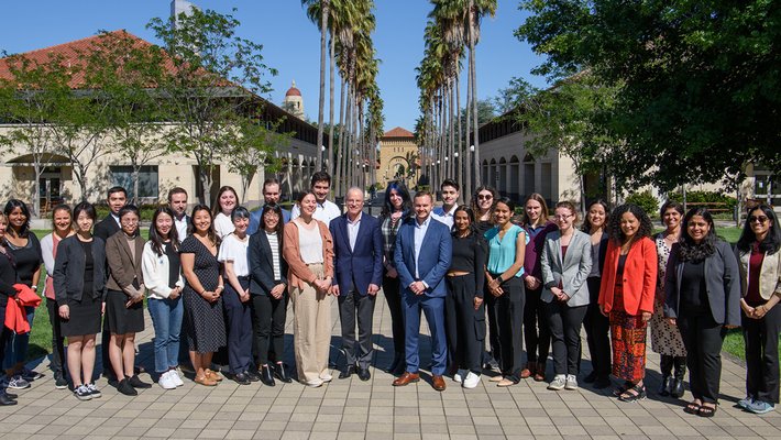 A group of scholars stand on a walkway in front of two rows of palm trees with Stanford buildings visible in the background.