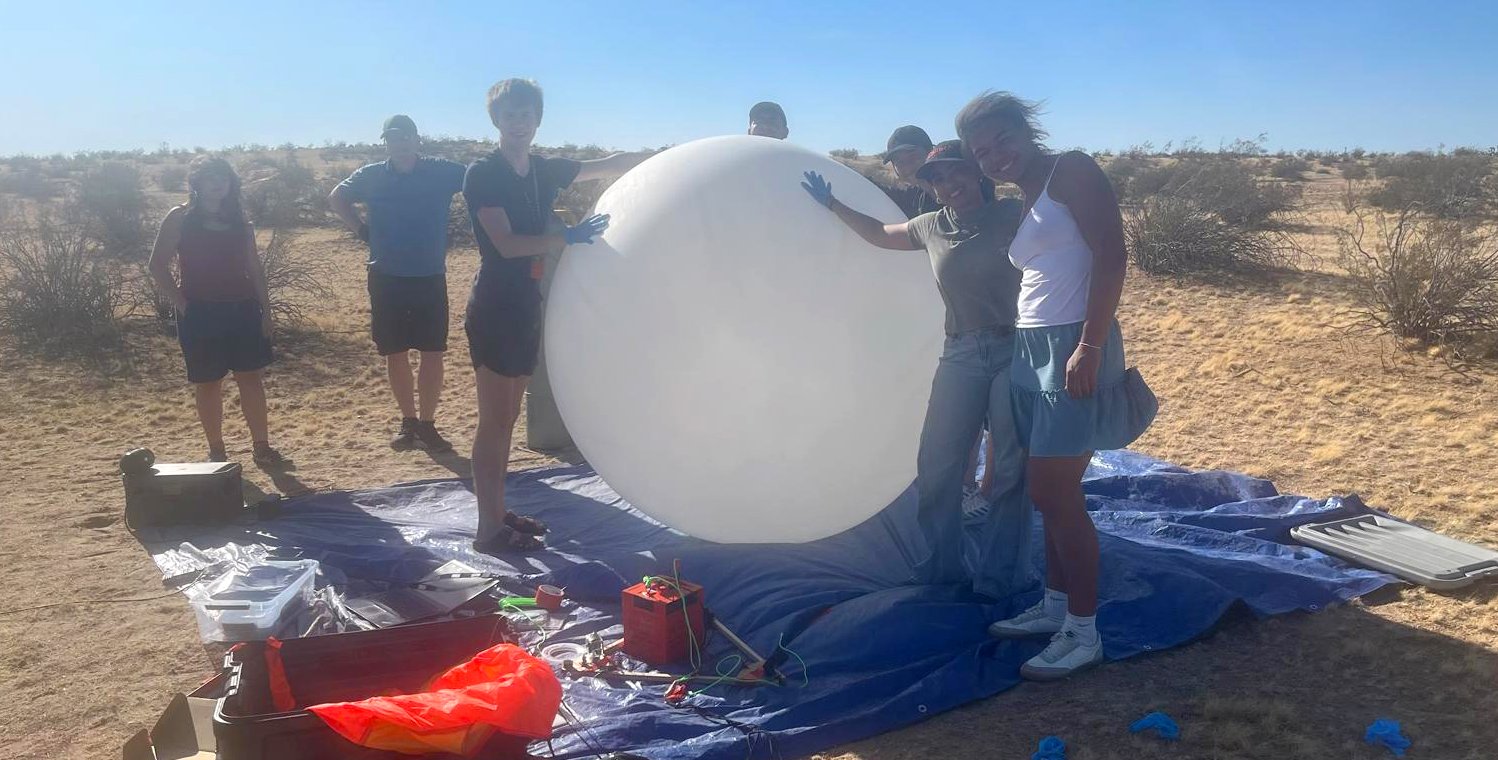 a mostly student group stands in the windy desert holding down a large, white helium-filled balloon over a blue tarp