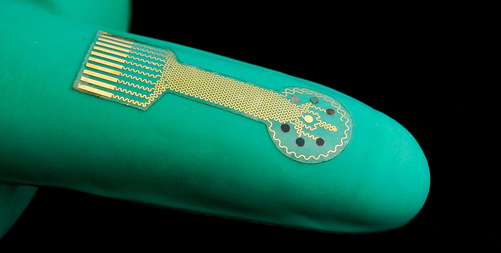A device consisting of a thin clear film with gold traces on it rests on top of a rubber-gloved finger.