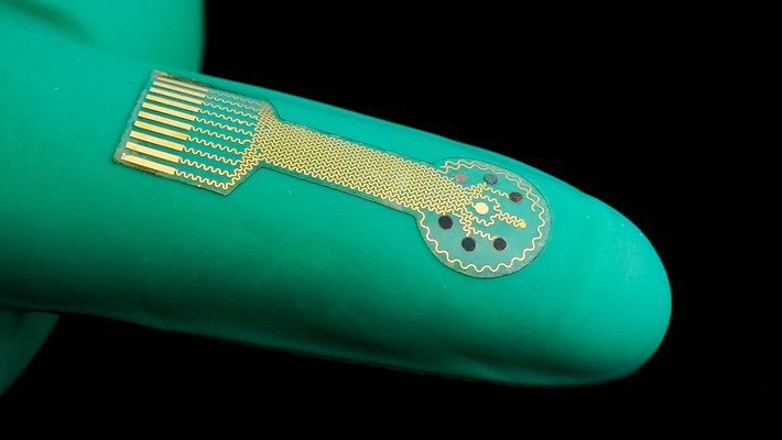 A device consisting of a thin clear film with gold traces on it rests on top of a rubber-gloved finger.