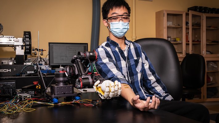 A human sits at a table flexing his hand. Sensors are attached to the skin of his forearm. A robotic hand next to him mimics his motion.