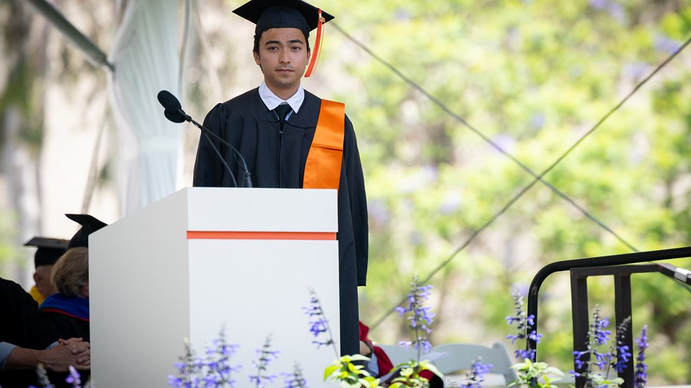 A young man wearing a black robe and mortarboard with an orange tassle stands as a commendation for him is read out.