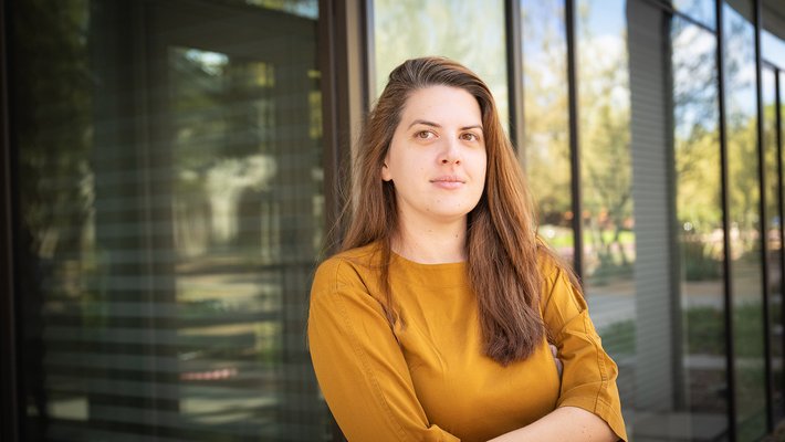 A woman with long auburn hair wearing a yellow, long-sleeved shirt poses with her arms crossed in front of a glass exterior wall.