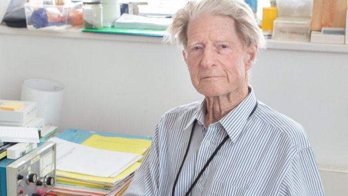 An older man sits at a desk wearing a blue pin-striped shirt.