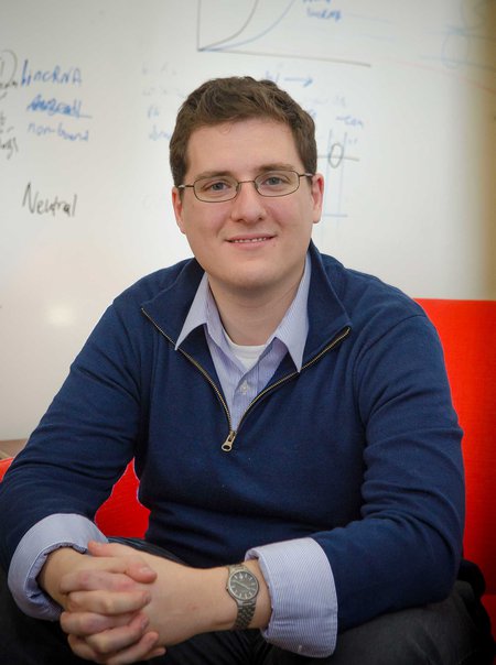 A man in glasses sits in front of a whiteboard and smiles.