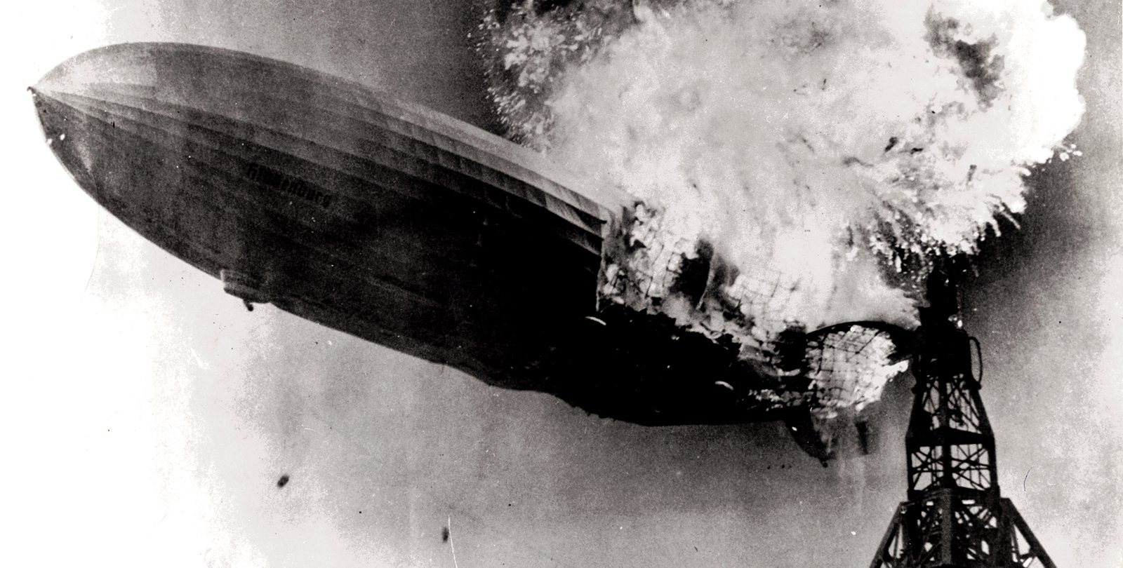 A large zeppelin, partially consumed by flames, begins sinking toward the ground next to its mooring mast. The image is a black and white photograph.