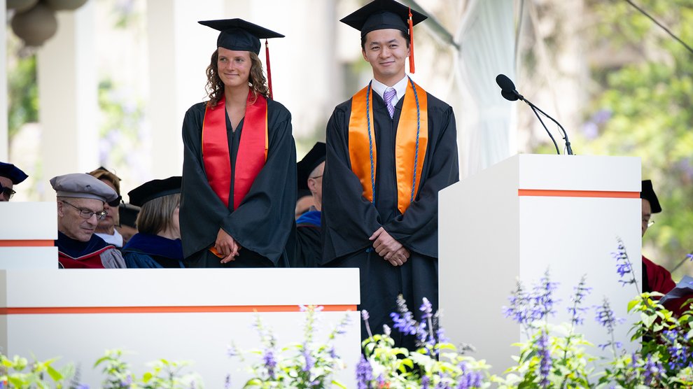 Two college graduates, both in graduation garb stand side by side. One is a young woman with a red sash and medium brown hair and the other is a young man with a lavender tie and short black hair.