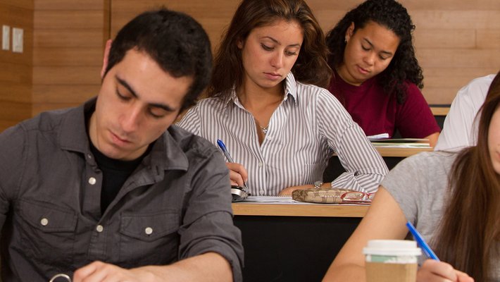 Five students photographed relatively close-up sit at long desks, writing with pen on paper.