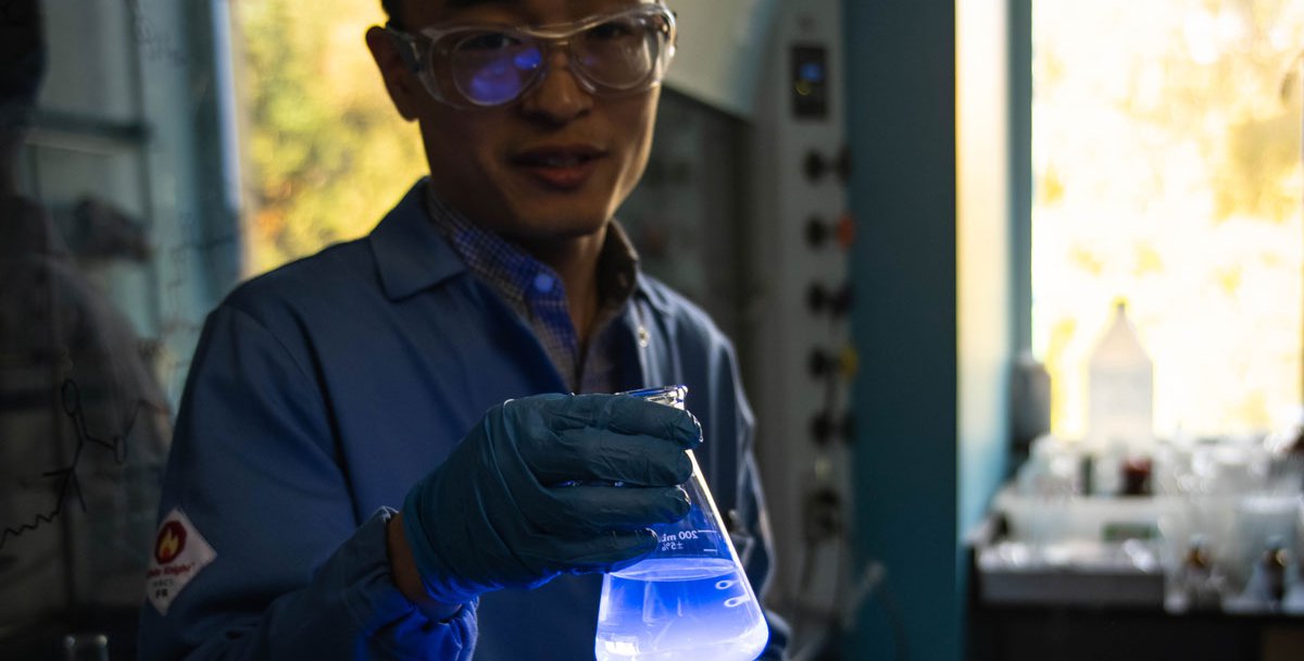 A researcher wearing a lab coat and safety glasses holds a flask of liquid in a dimly-lit lab. The flask glows a bright blue color.