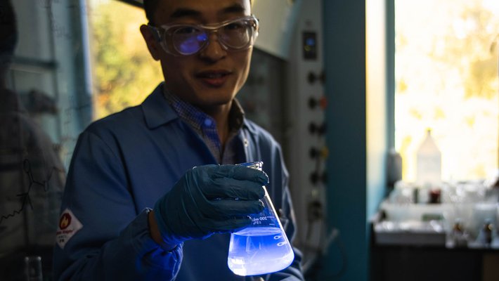 A researcher wearing a lab coat and safety glasses holds a flask of liquid in a dimly-lit lab. The flask glows a bright blue color.