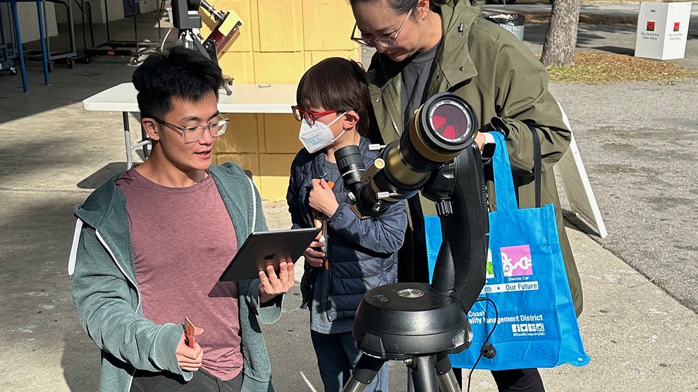 Three people gather behind a telescope on a sunny day