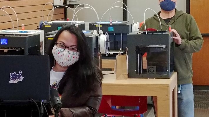A female in the foreground and male in the background, both at 3D printers and wearing masks