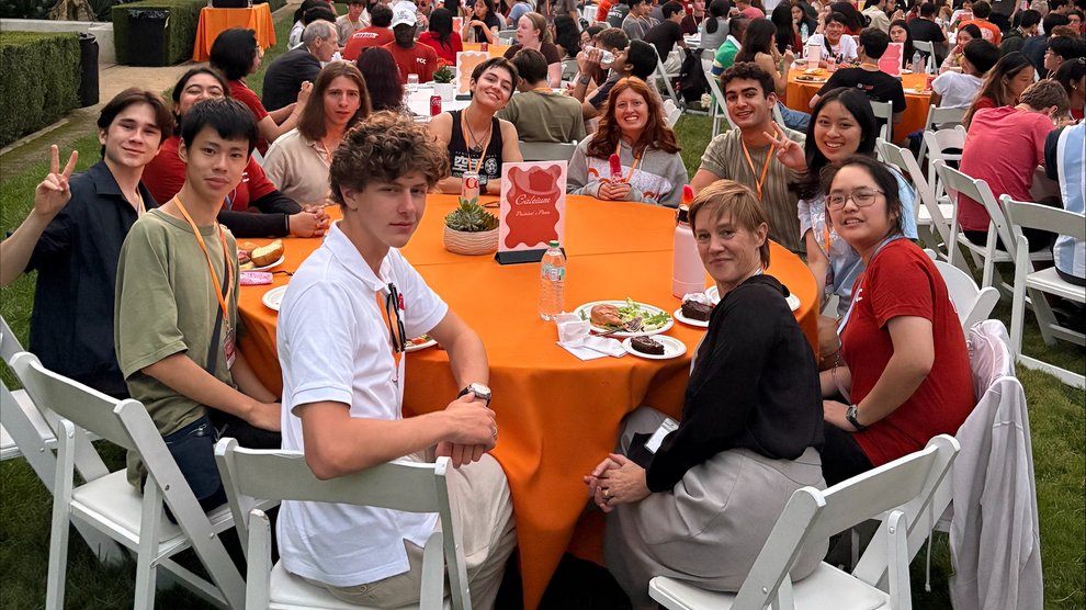 Incoming first-years sit at a table with Dean Jennifer Jahner and pose in front of a pink and orange sunset.