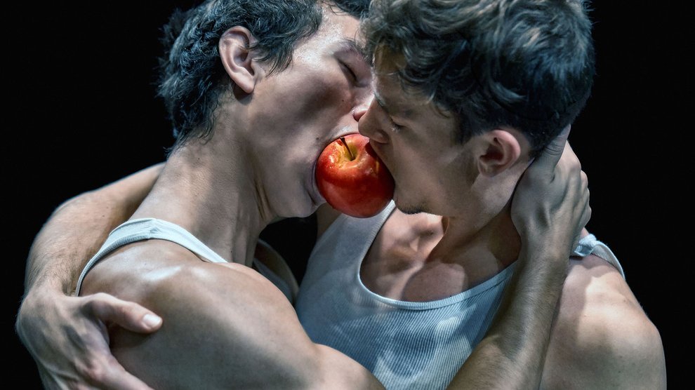 Two men embrace one another with an apple in between their mouths