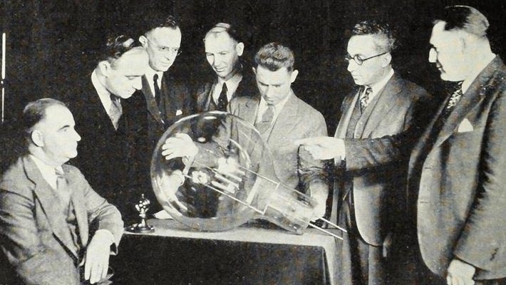 A black and white photograph from 1929 of seven men in suits, one seated and six standing, looking at an electric lightbulb the size of a beach ball resting on a table before them.