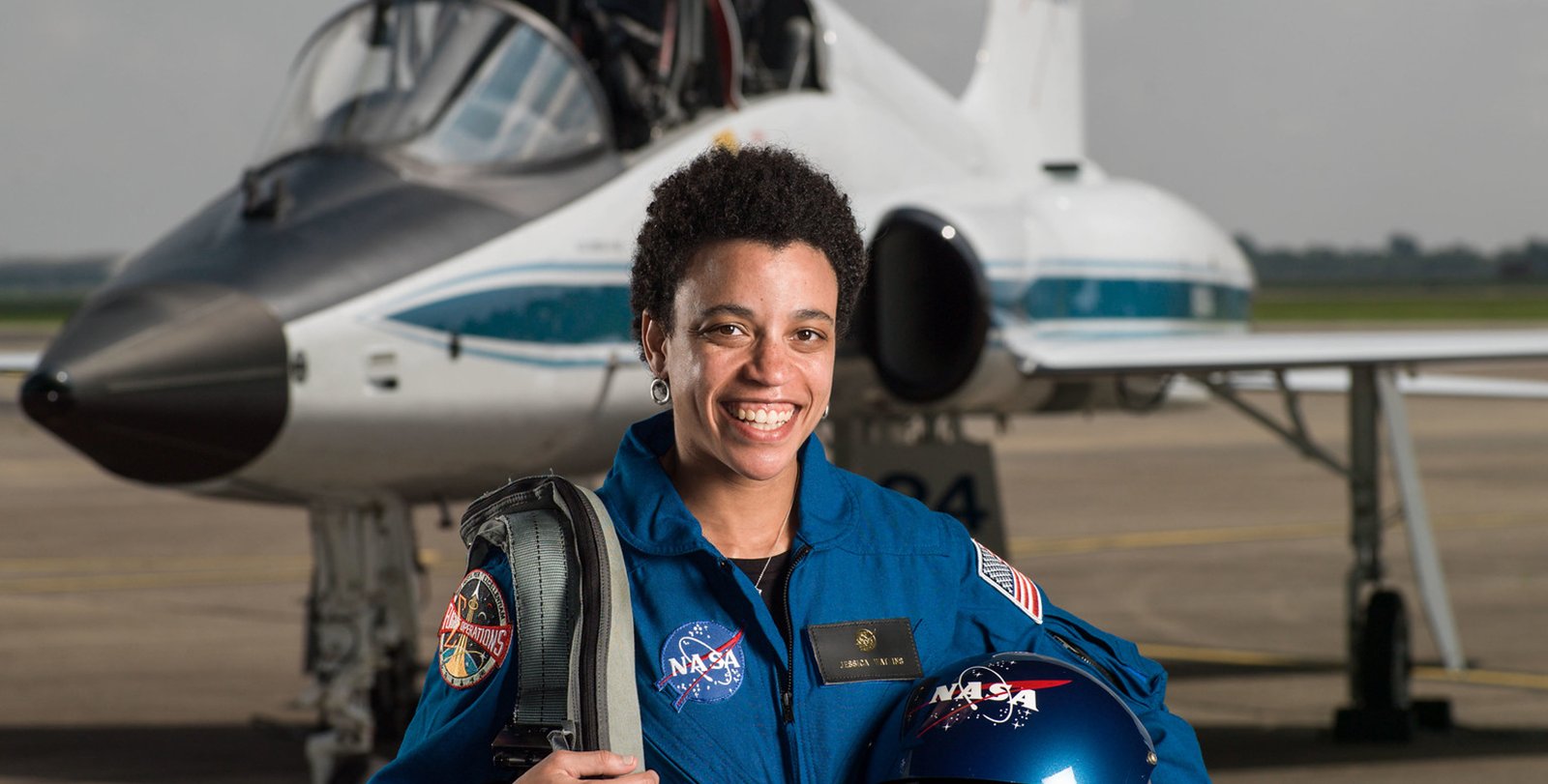 Jessica Watkins, in a blue flight suit and carrying a helmet, stands in front of a T-38 trainer aircraft