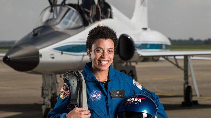 Jessica Watkins, in a blue flight suit and carrying a helmet, stands in front of a T-38 trainer aircraft