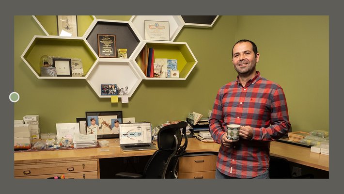 Joe Parker in his office, wearing a flannel shirt and holding a coffee cup next to hexagonal shelving