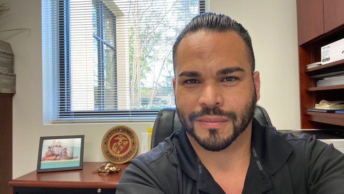 Selfie of Juan Balcazar wearing a Caltech polo in his office with a Marine Corp sign and photo of a family in the background