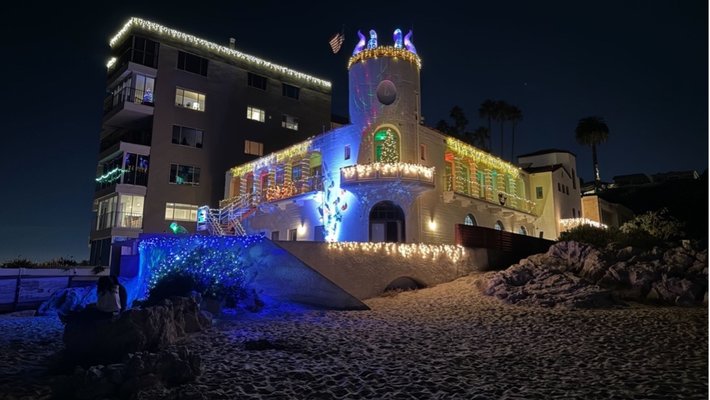 A large stucco building decorated with holiday lights stands above a beach, in front of decorated condominiums