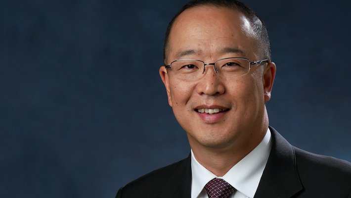 Portrait of David Kang wearing glasses and a dark suit and tie.